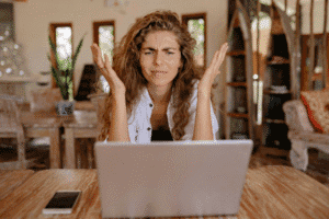 Confused woman with curly hair sitting at a wooden table using a laptop with a smartphone nearby.