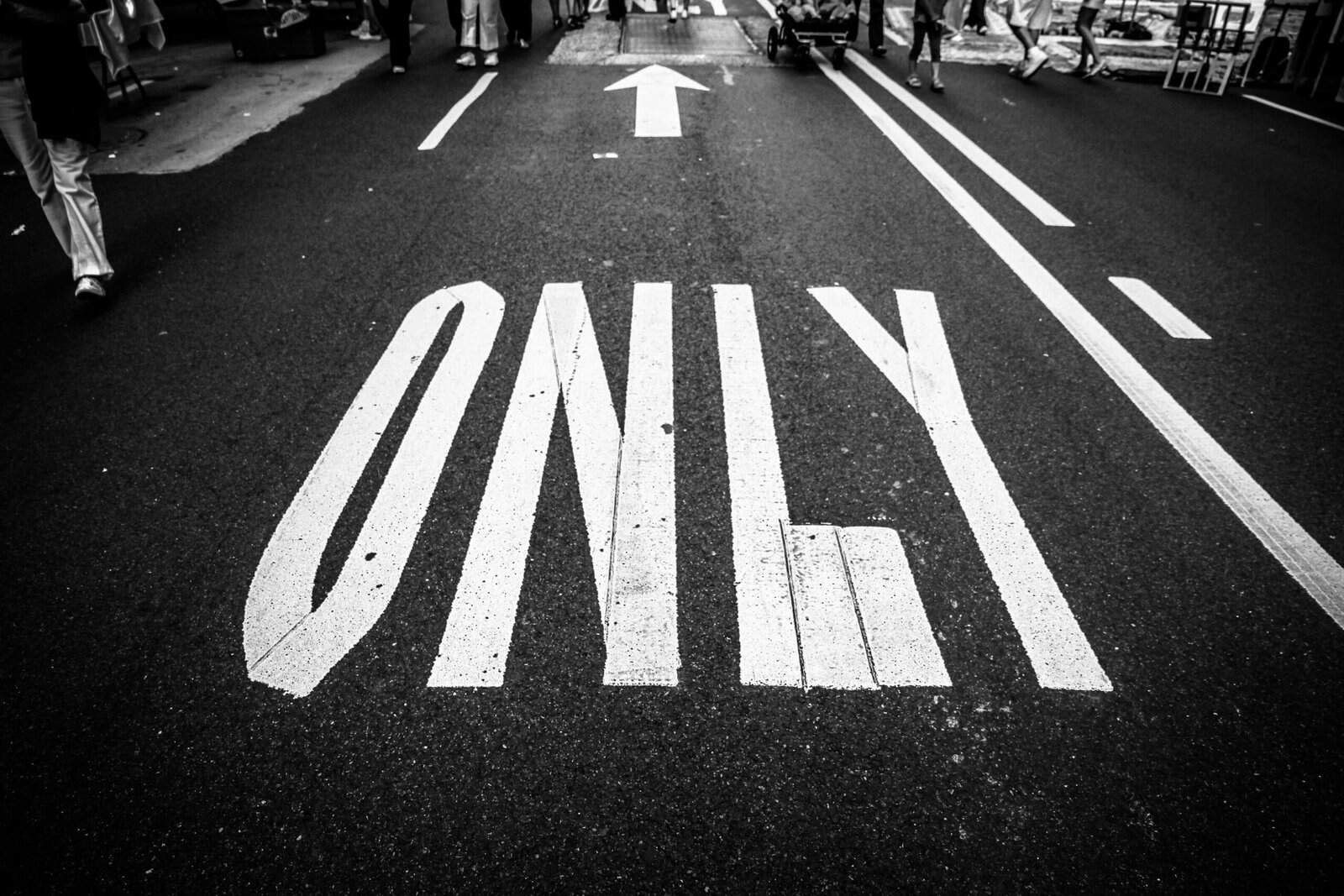 Black and white image of a painted stop sign on asphalt road, with pedestrians walking nearby.