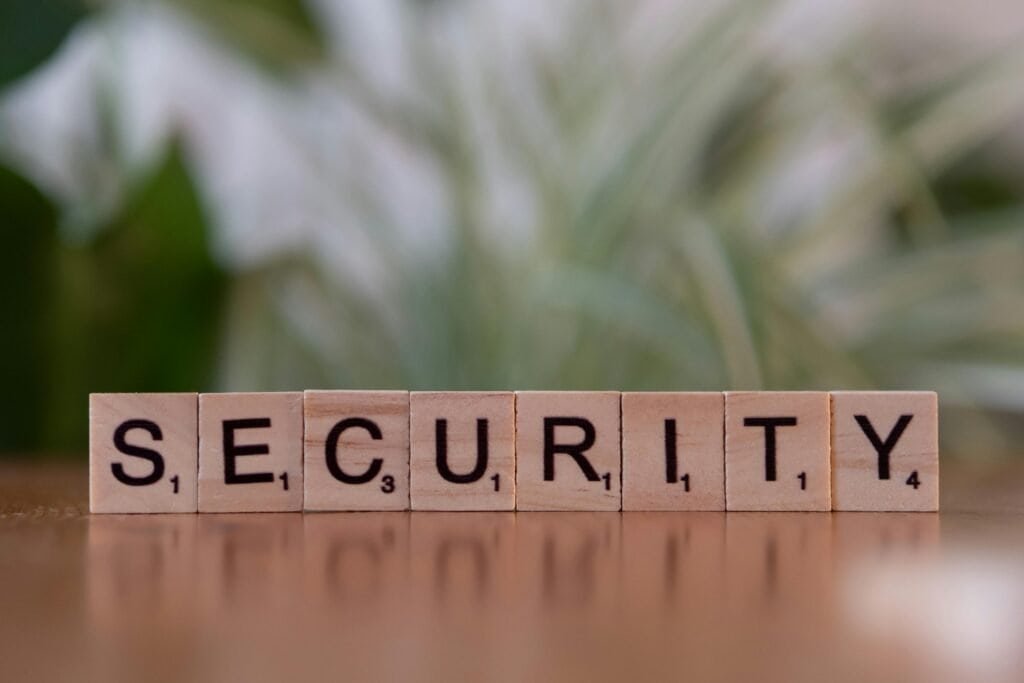 Close-up of wooden letter tiles spelling "SECURITY" on a wooden surface. Background is softly blurred greenery.